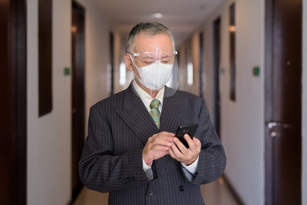 Mature Japanese businessman with mask and face shield using phone in the corridor